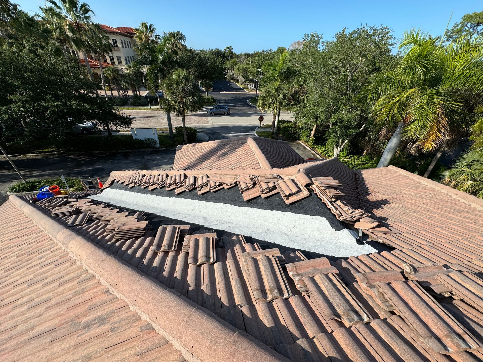 Rooftop with red tiles undergoing repair; silver flashing exposed, surrounded by broken tiles; palm trees in background. Rooftop with red tiles undergoing repair; silver flashing exposed, surrounded by broken tiles; palm trees in background.