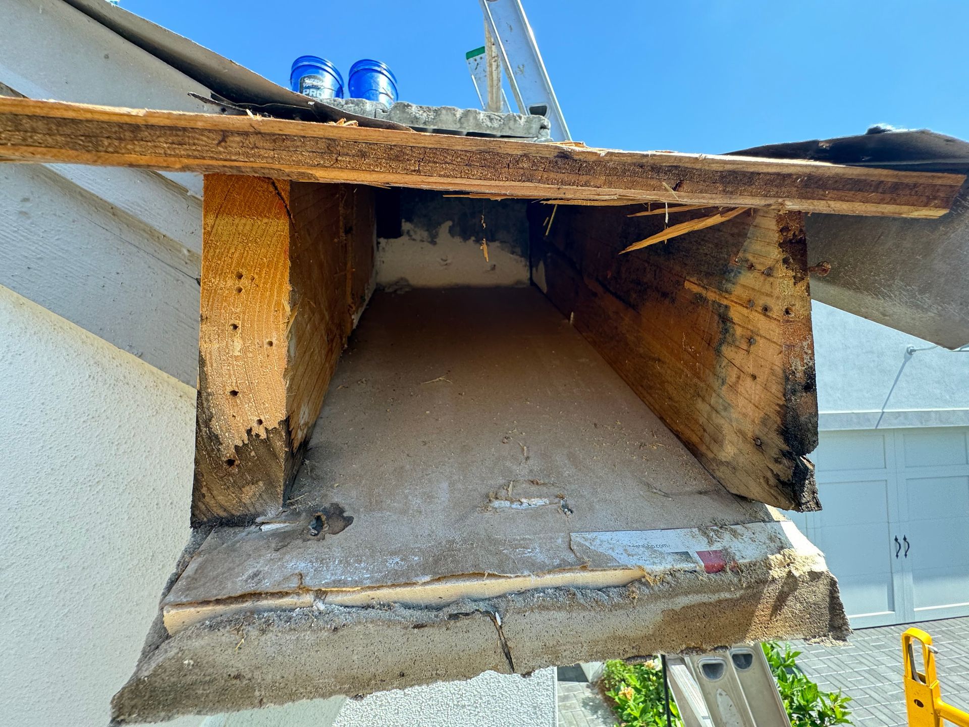 Damaged roof overhang showing exposed wood and possible water damage. Blue containers visible on the roof.