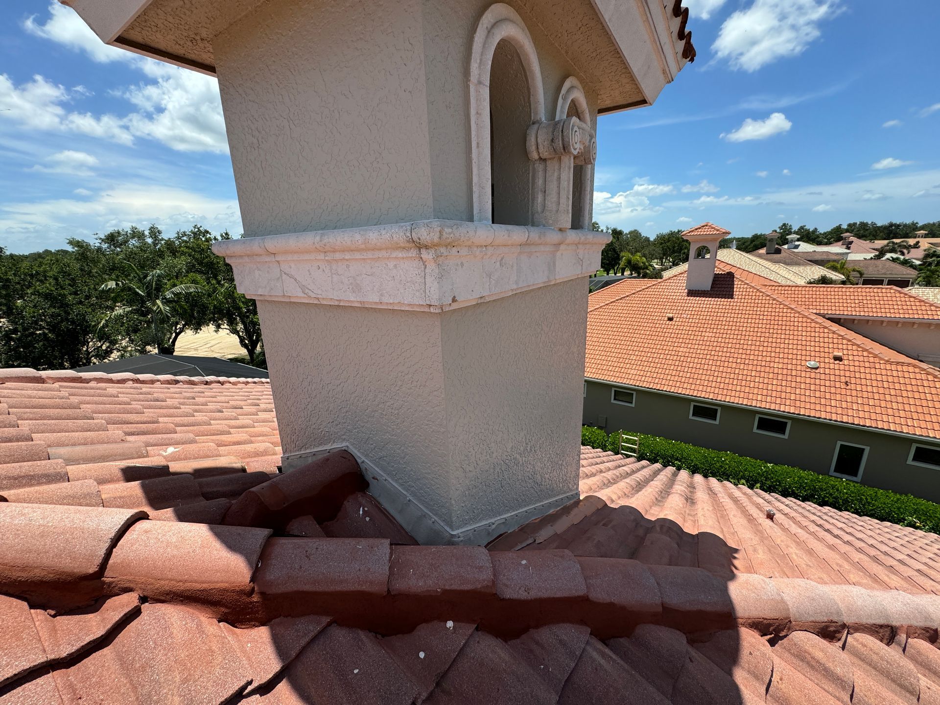 Chimney on a red tile roof, against a blue sky, with visible surrounding houses.