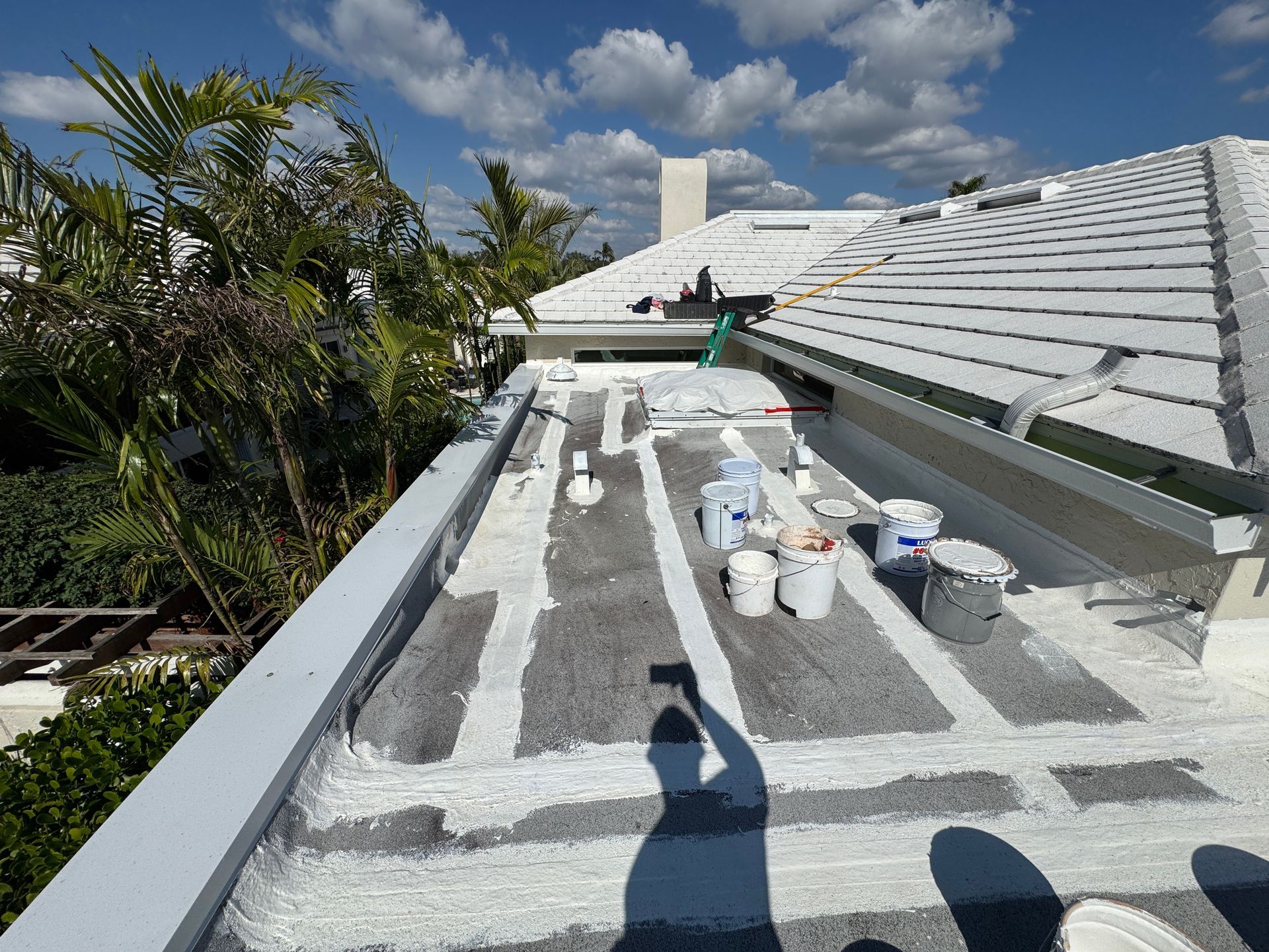 Flat roof being worked on, with white sealant applied in strips. Buckets and tools on the roof under a blue sky. Flat roof being worked on, with white sealant applied in strips. Buckets and tools on the roof under a blue sky.