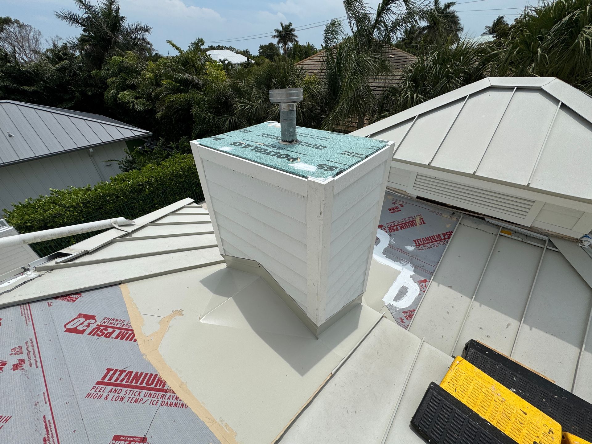 White chimney on a metal roof, with new roofing materials in place, surrounded by trees.