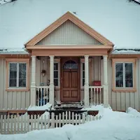 Snow-covered beige house with a wooden porch and peaked roof in winter