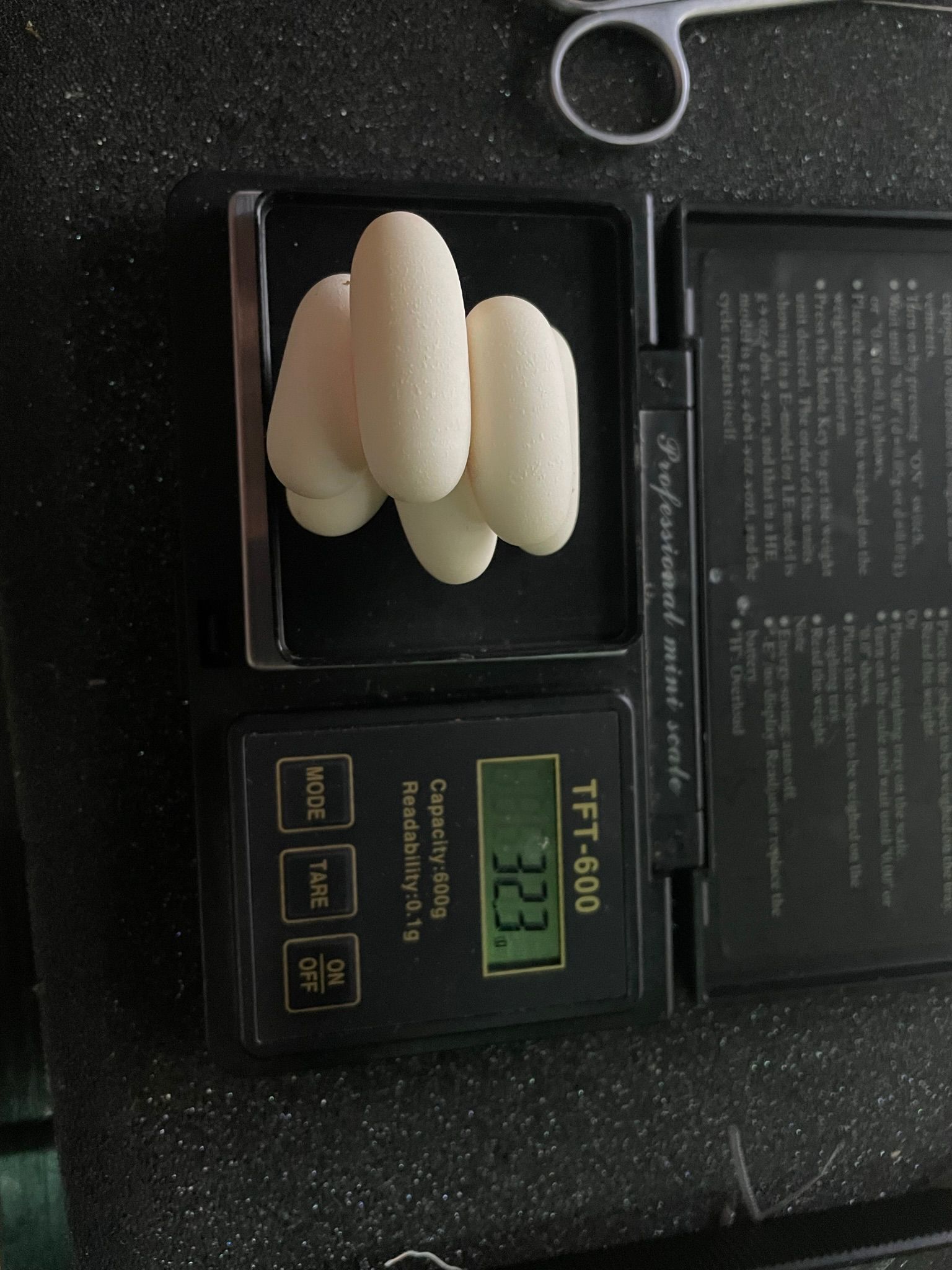 Weiße, längliche Tabletten auf einer Digitalwaage, deren Display 23,3 Gramm anzeigt, und eine Schere daneben.
