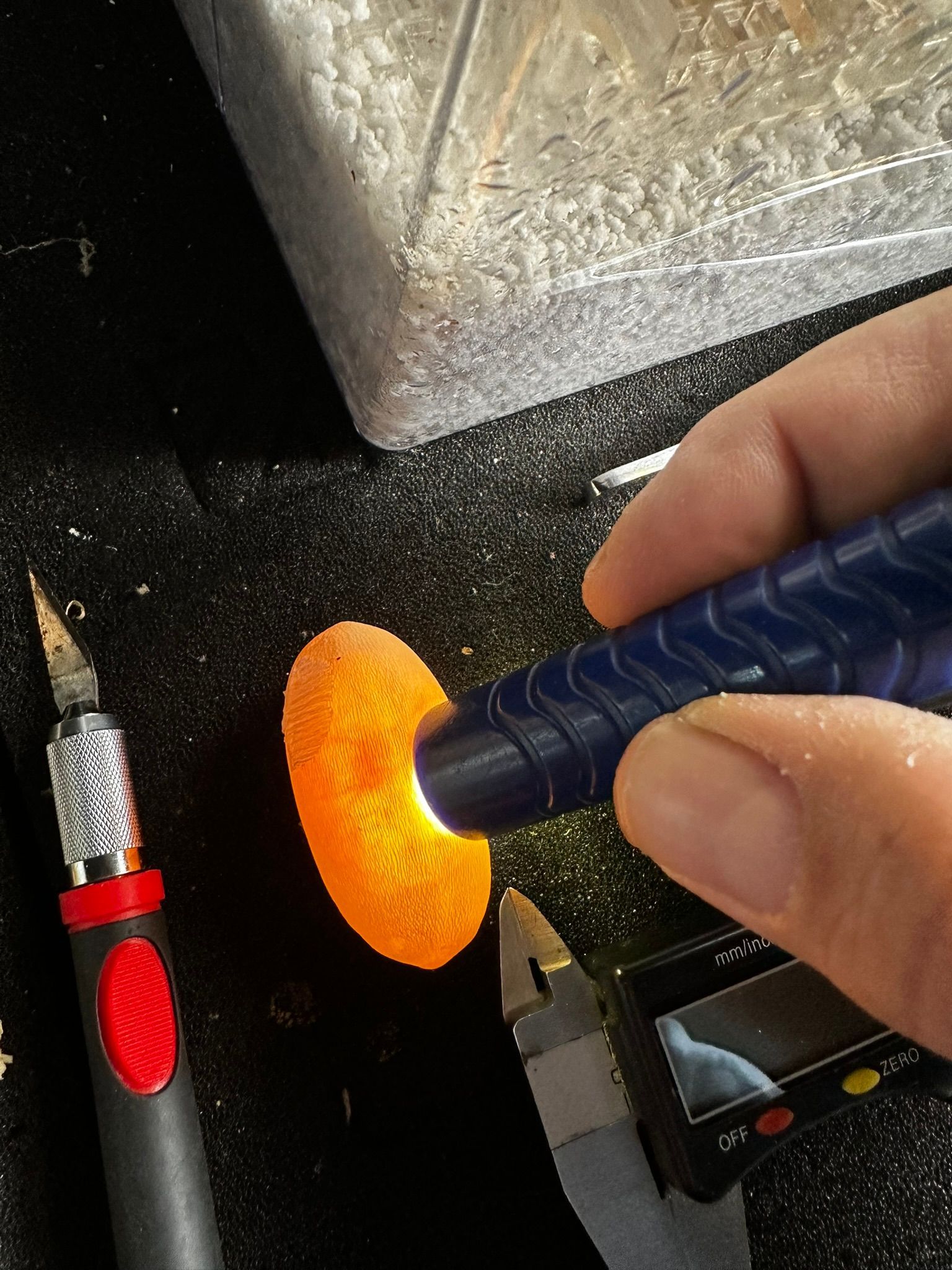 Eine Hand hält eine blaue Taschenlampe auf ein leuchtend orangefarbenes Metallstück auf einer dunklen Werkbank.