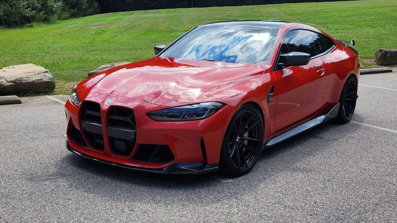 Red BMW M4 coupe parked on asphalt, green grass and a rock in the background. Black wheels.