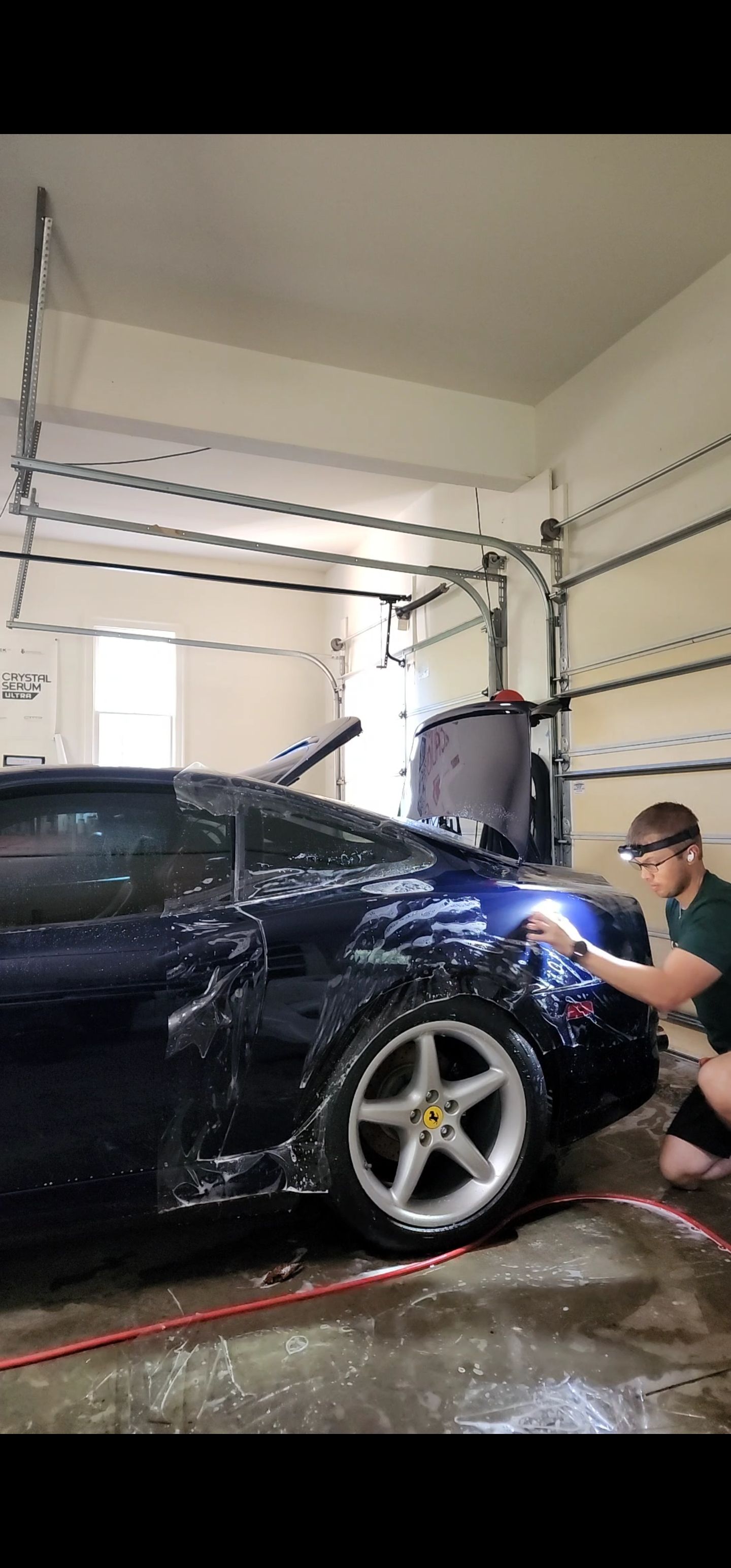 Man washing a black sports car in a garage, spraying water near the front wheel