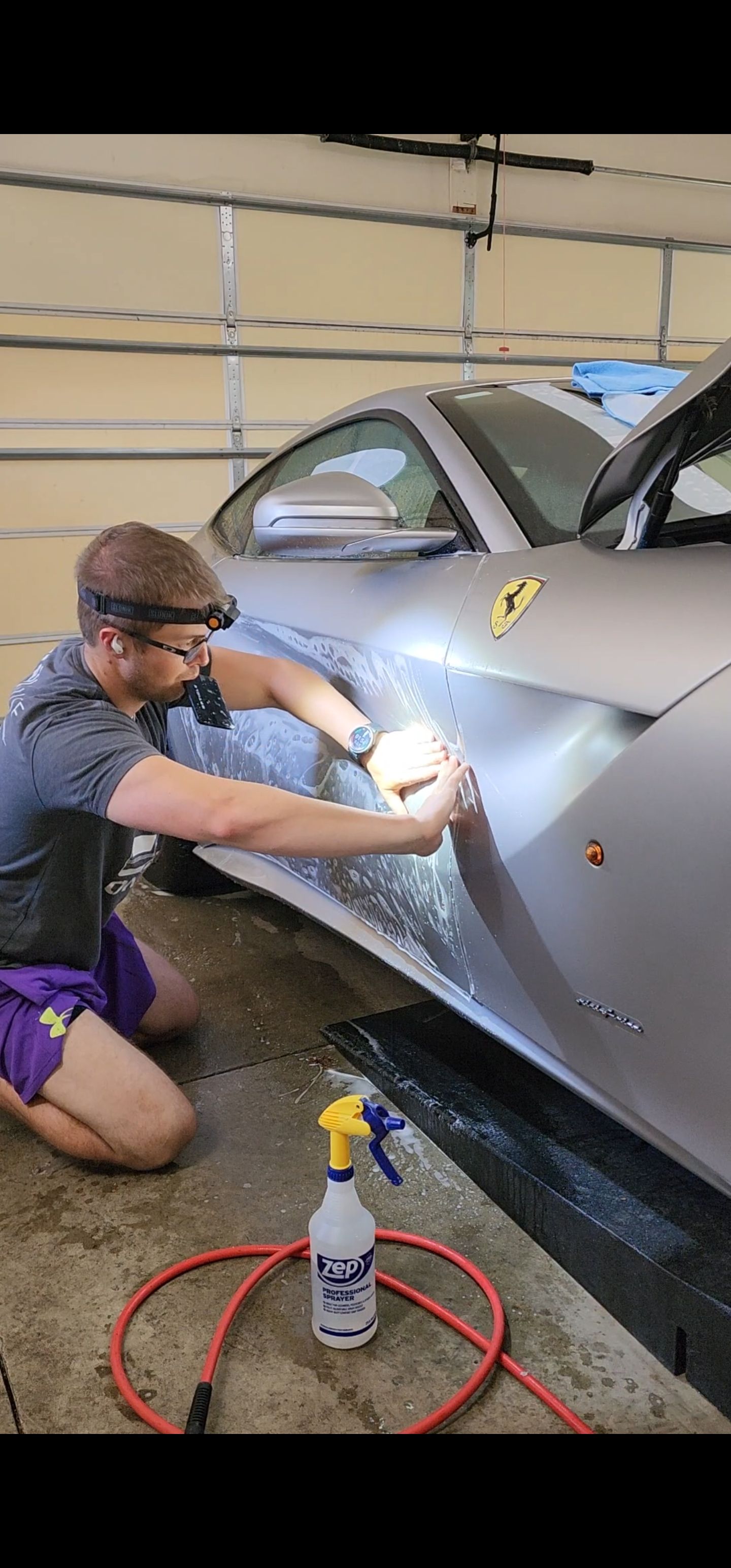 Mechanic polishing a silver sports car in a garage with a spray bottle on the floor