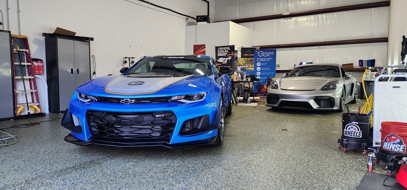 Blue Camaro and silver sports car in a garage with tools and equipment.
