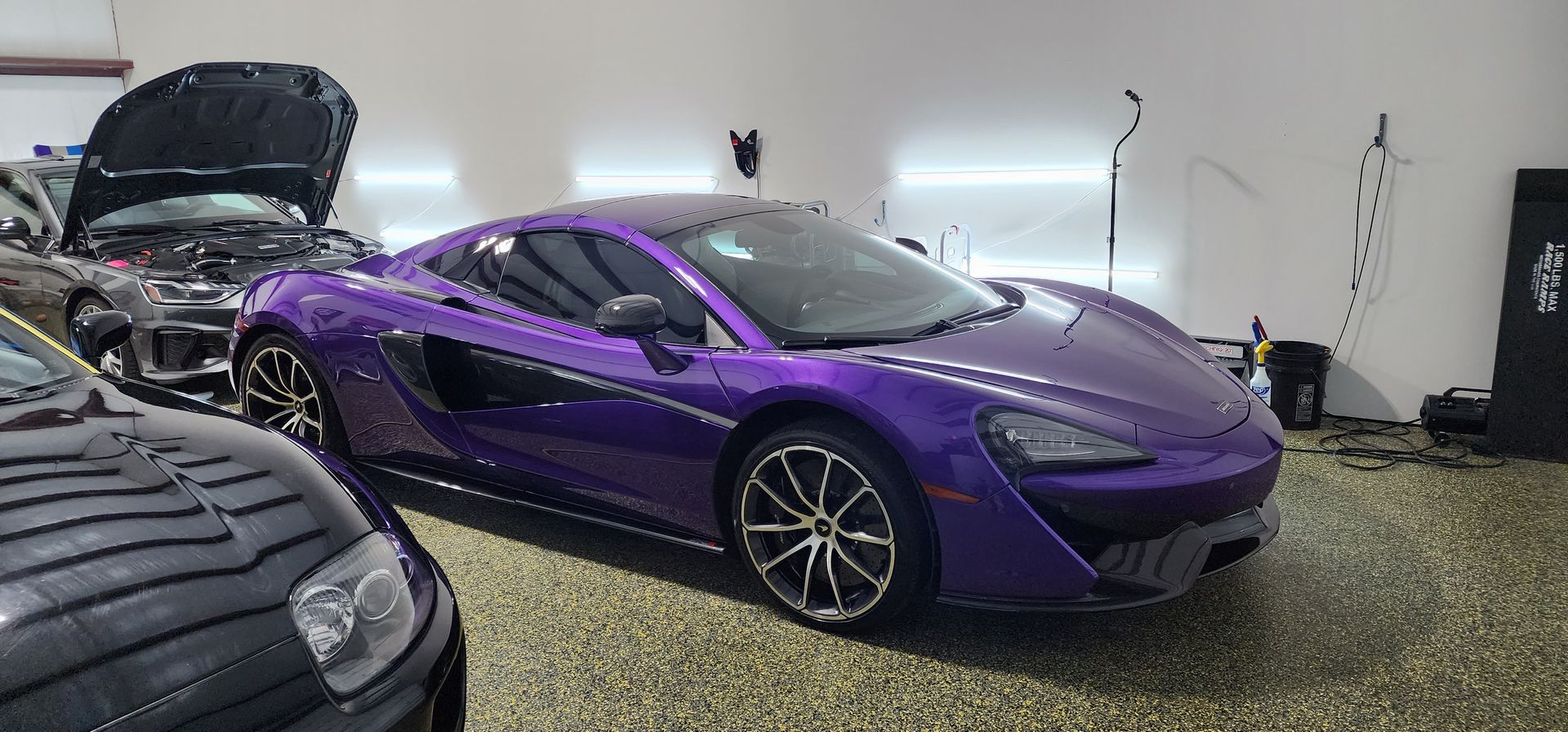 A purple McLaren sports car in a garage. Another car is in front, with hood open.