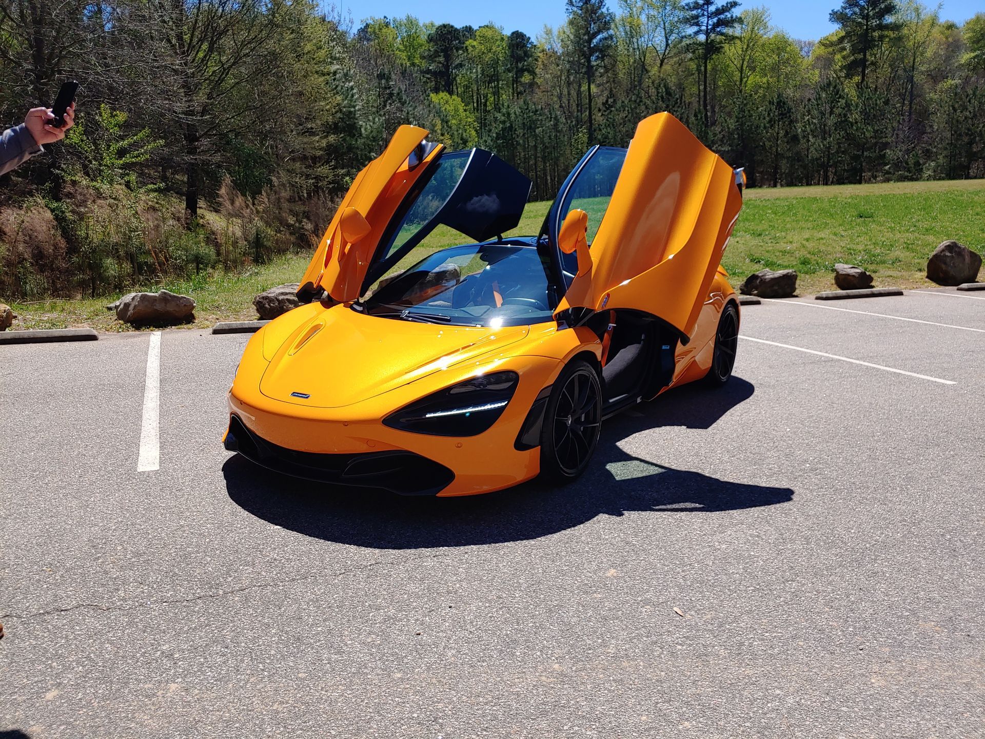Orange sports car with butterfly doors open in a parking lot, trees in the background