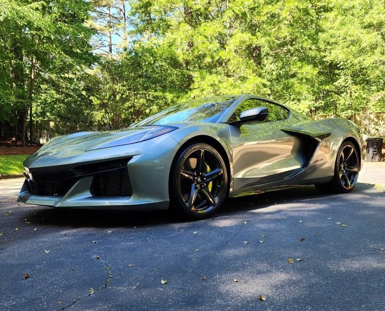 Silver sports car parked on a sunlit road beside green trees.