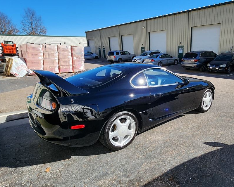 Black Toyota Supra parked in a lot, with other cars and a building in the background. Sunny day.