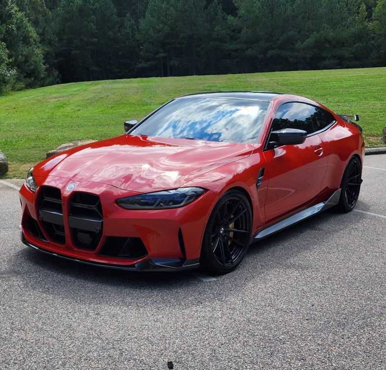 Red sports sedan parked on pavement, with a grassy field and trees in the background.