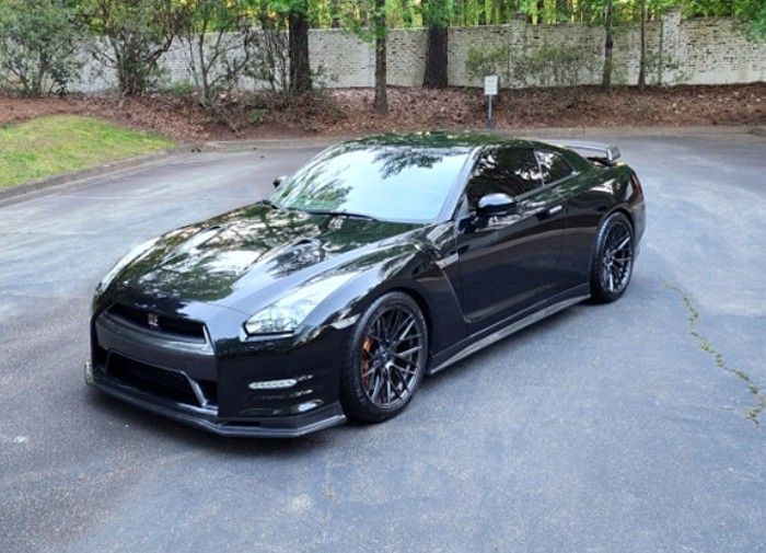 Black sports coupe parked on a paved driveway with trees and stone wall in the background