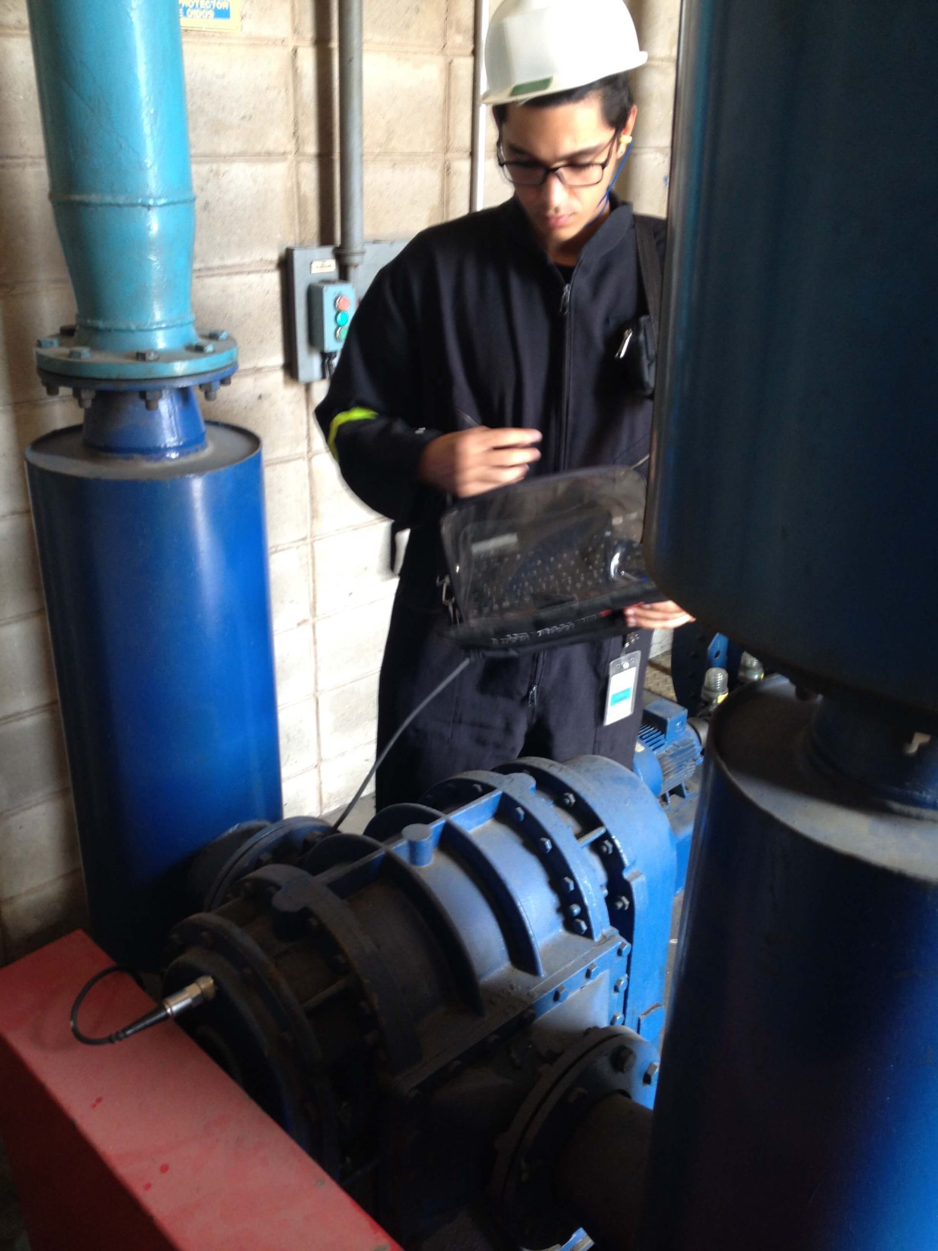 Una persona con un mono azul y casco utiliza una computadora portátil cerca de una maquinaria industrial azul.