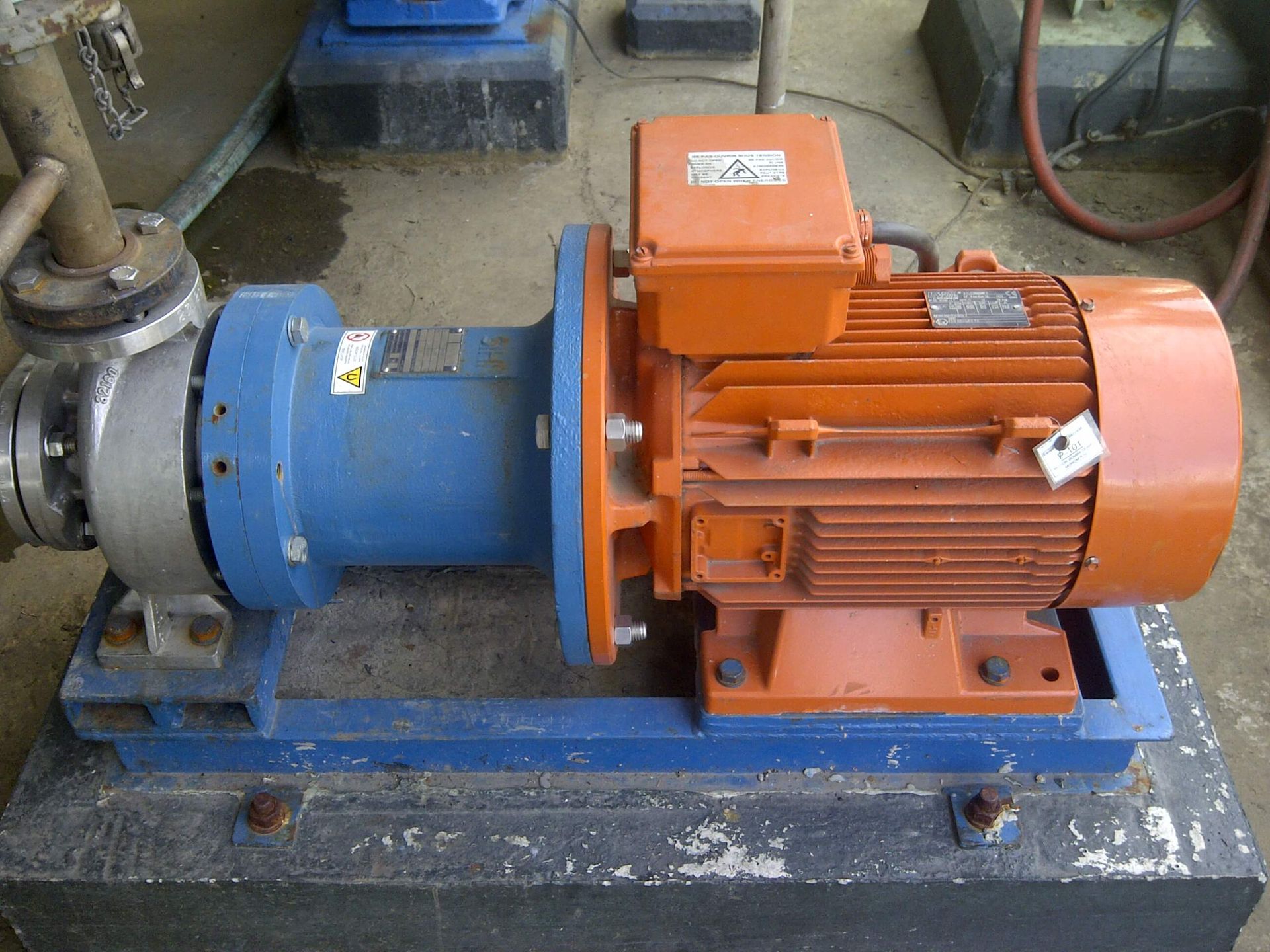 Una bomba industrial azul y naranja sobre una base de hormigón, conectada a una tubería y alimentada por un motor eléctrico.