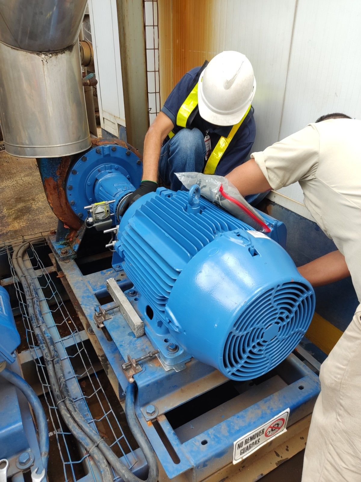 Dos trabajadores con chalecos de seguridad y cascos reparan un gran motor azul en un entorno industrial.