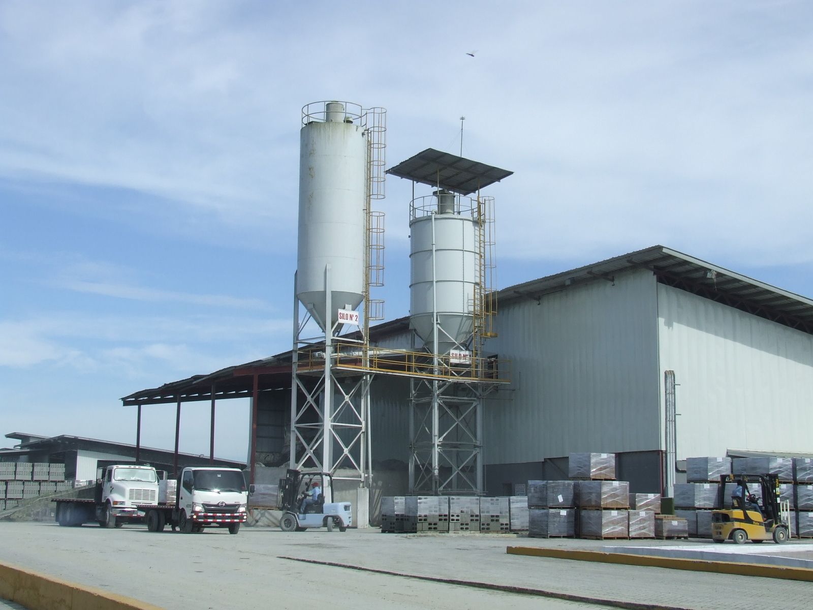 Fábrica con silos, camiones y carretillas elevadoras, con un cielo azul.