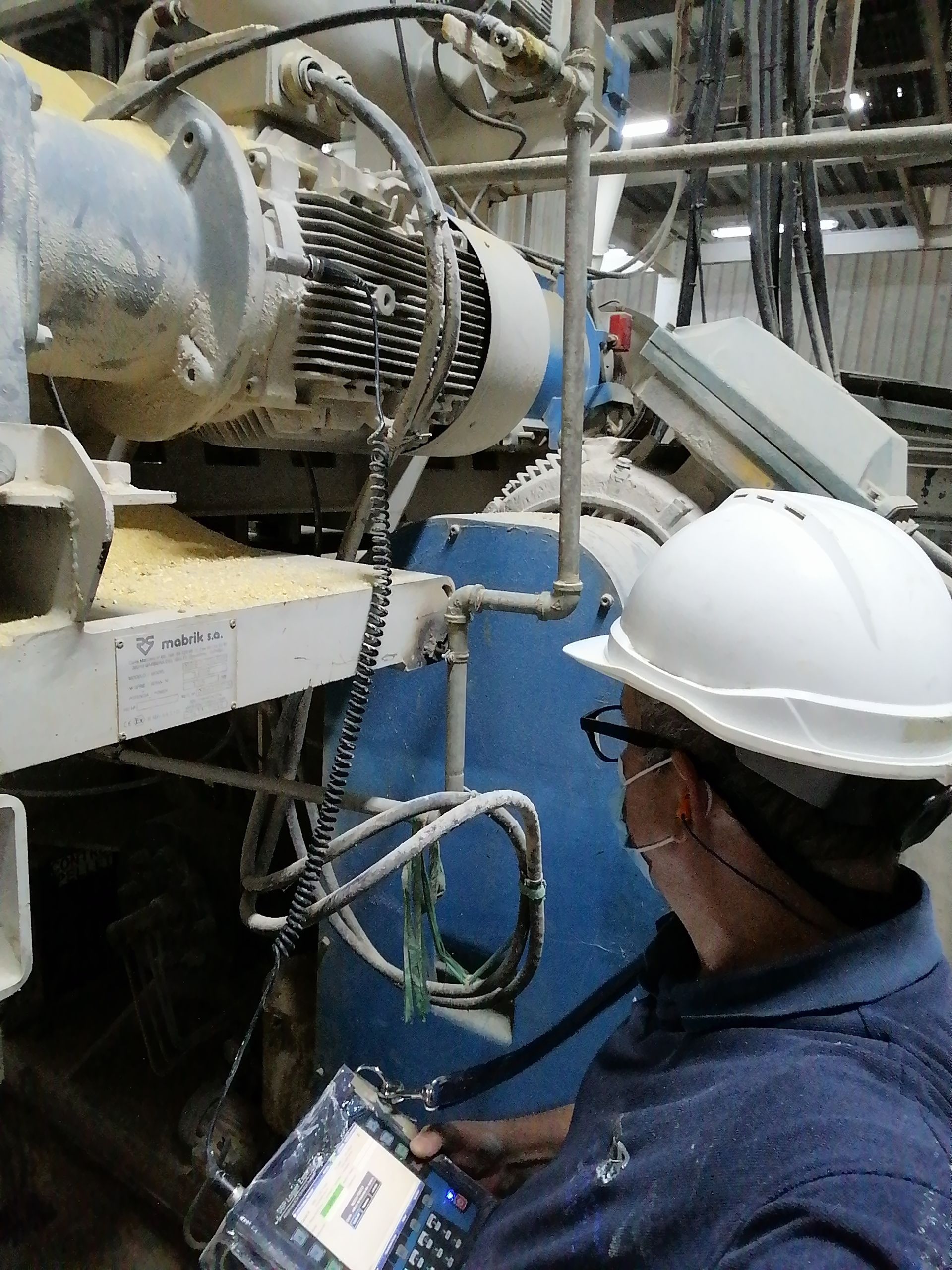 Una persona con casco inspecciona maquinaria con un dispositivo portátil. Equipo industrial azul y gris.