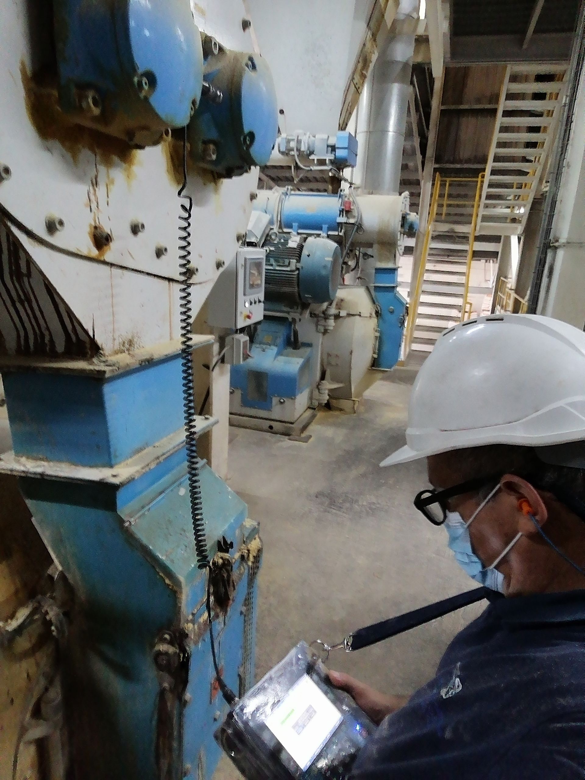 Una persona con casco y máscara utiliza un dispositivo en un entorno industrial con maquinaria y una escalera.