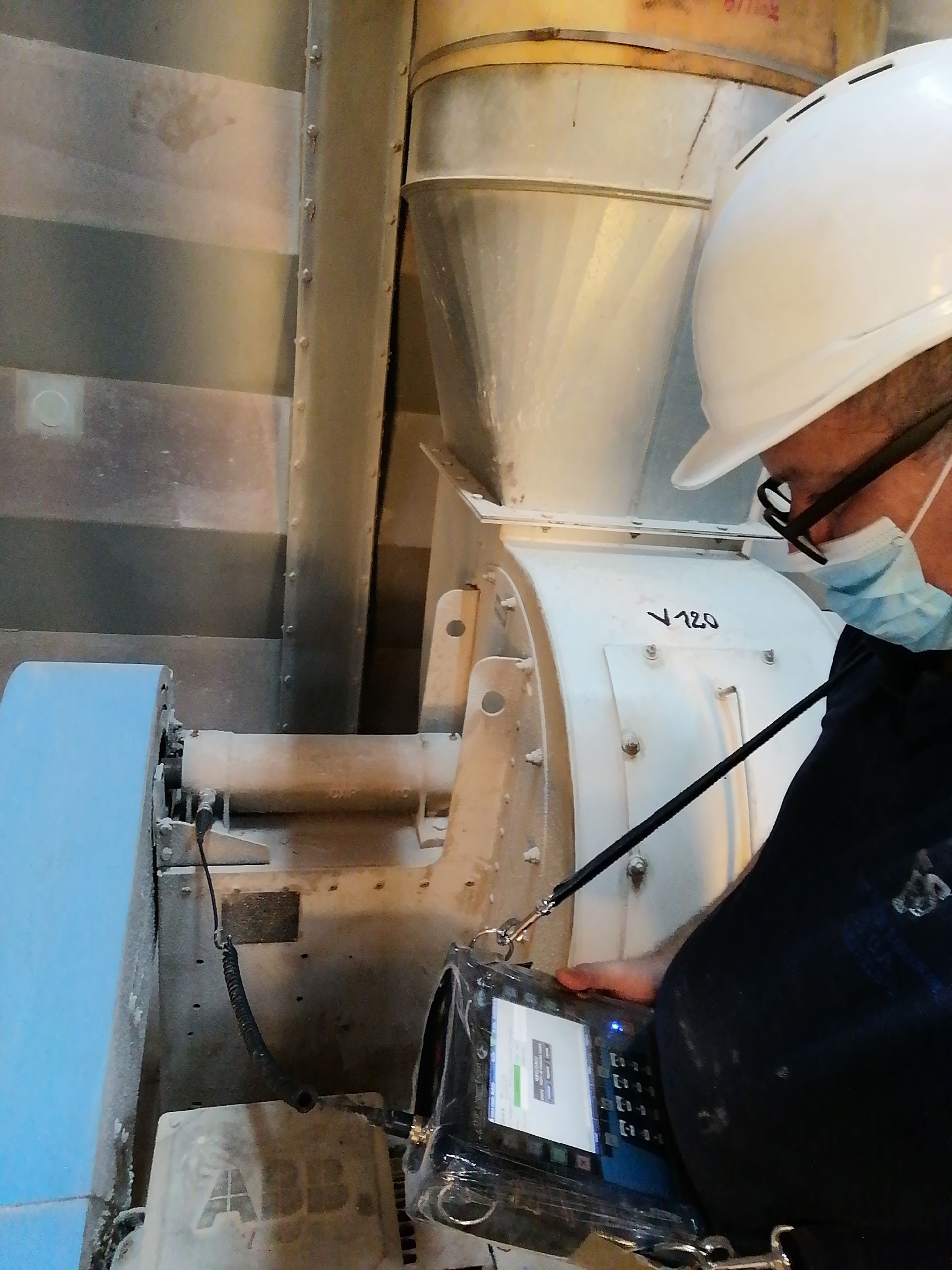 Una persona con casco y máscara utiliza un dispositivo portátil para inspeccionar maquinaria industrial.