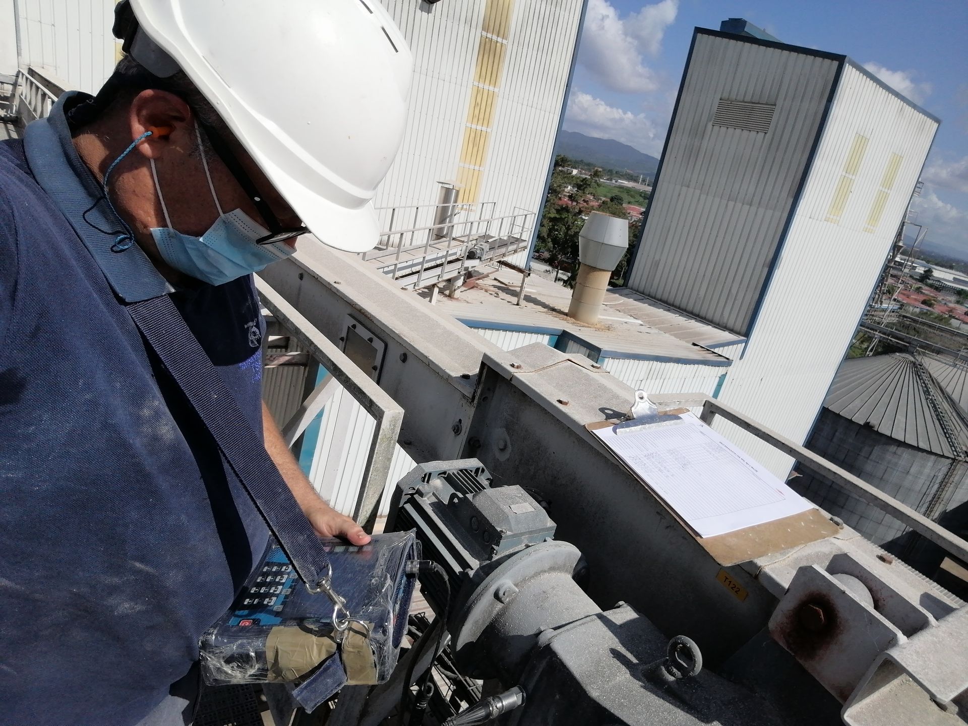 Persona con casco y mascarilla examinando maquinaria en una azotea. Edificios y cielo al fondo.