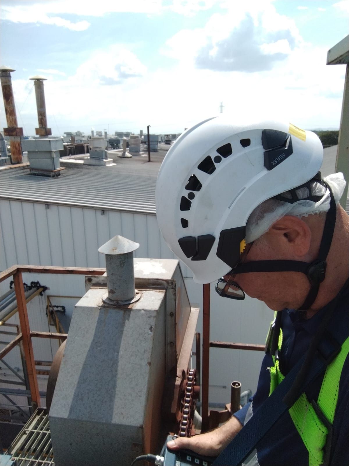 Persona con casco blanco y chaleco de seguridad en una azotea, inspeccionando equipo.