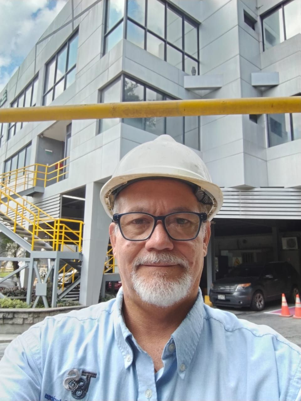 Hombre con casco y gafas frente a un edificio industrial.