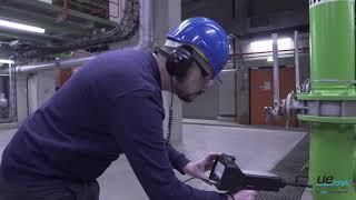Hombre con casco azul y protección auditiva inspeccionando maquinaria con un dispositivo portátil en una fábrica.