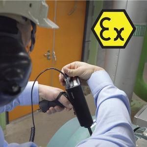Trabajador usando una herramienta cerca de maquinaria. Marca EX en hexágono amarillo.