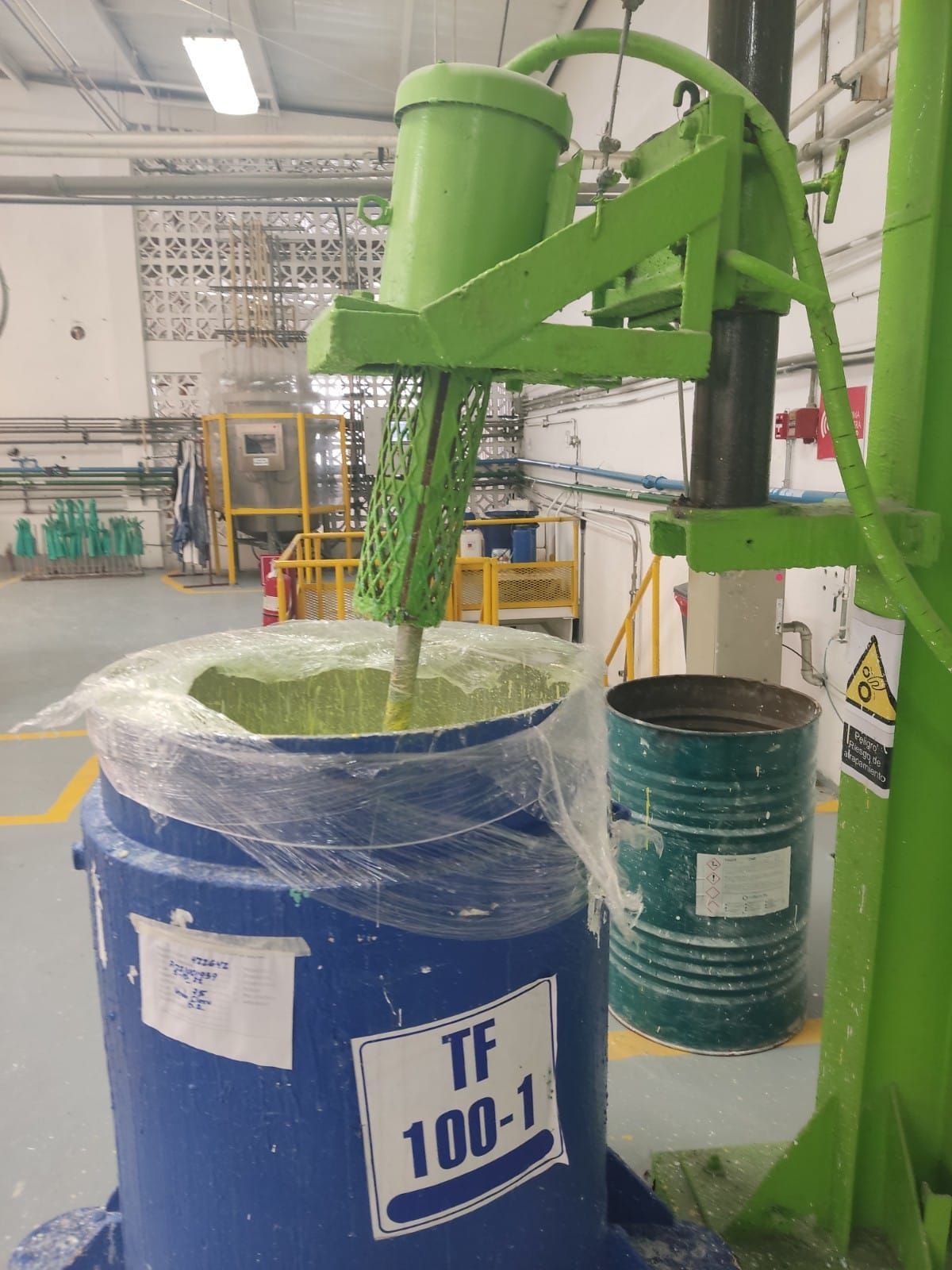 Mezclador industrial verde que revuelve una sustancia verde dentro de un barril azul en un entorno de fábrica.