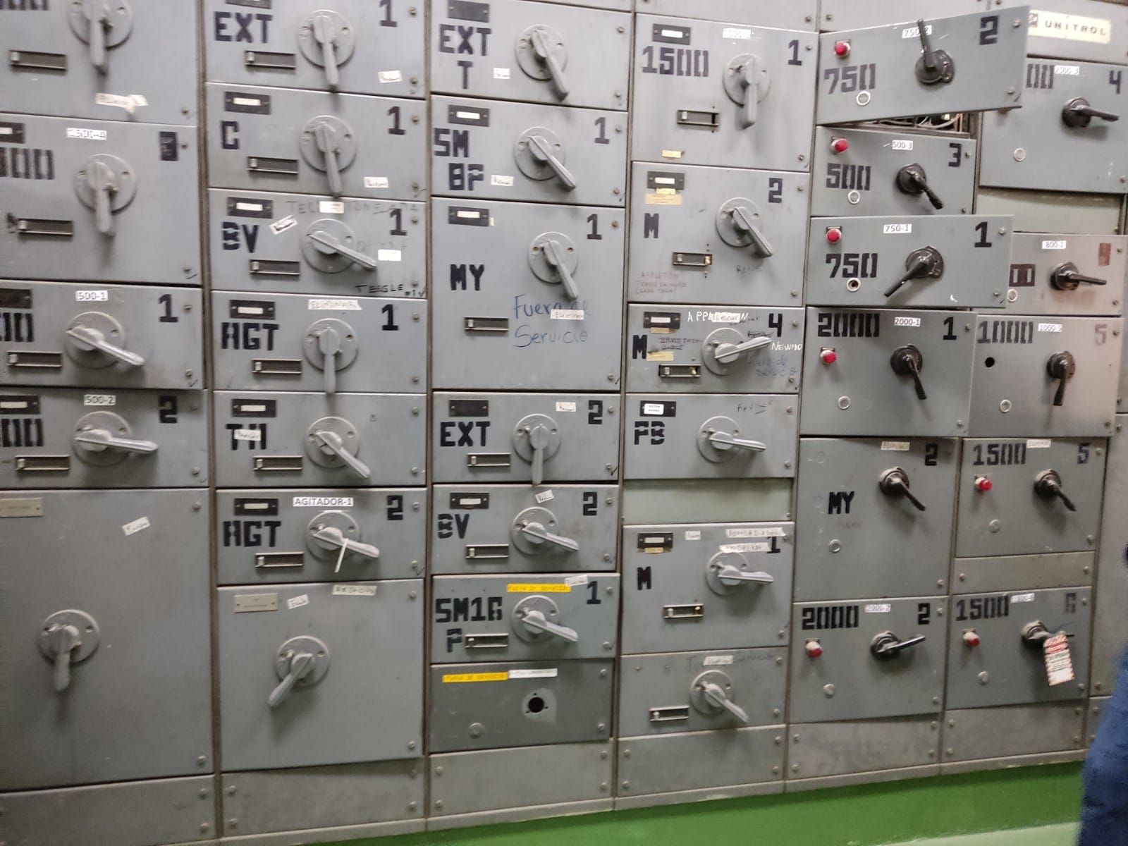 Panel de cajas eléctricas grises con interruptores, etiquetados con números y abreviaturas.