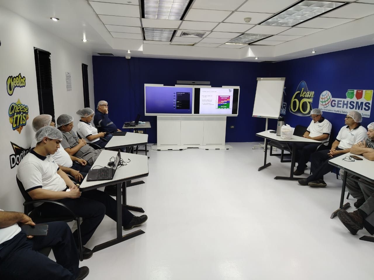 Grupo de personas con redecillas en una sala de formación con mamparas y mesas.