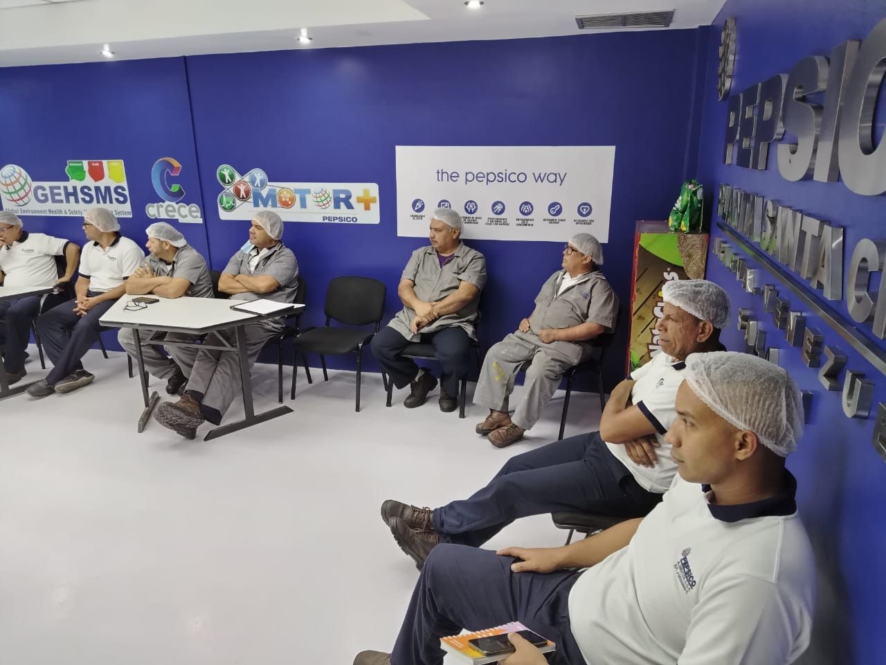 Un grupo de personas con redecillas, en una sala de paredes azules, parece estar reunido. Algunos están sentados, escuchando.