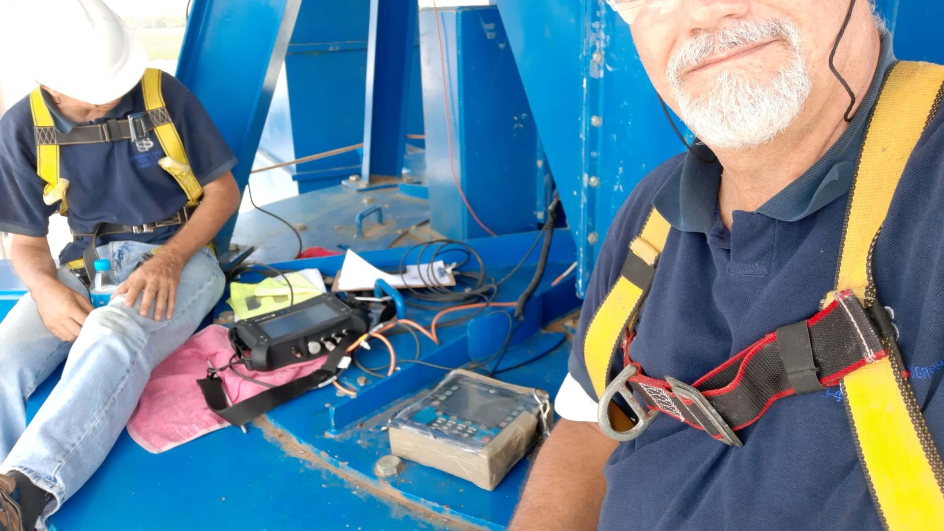 Dos personas con arneses de seguridad en una estructura azul, con equipo cerca. Una persona se toma una selfie.