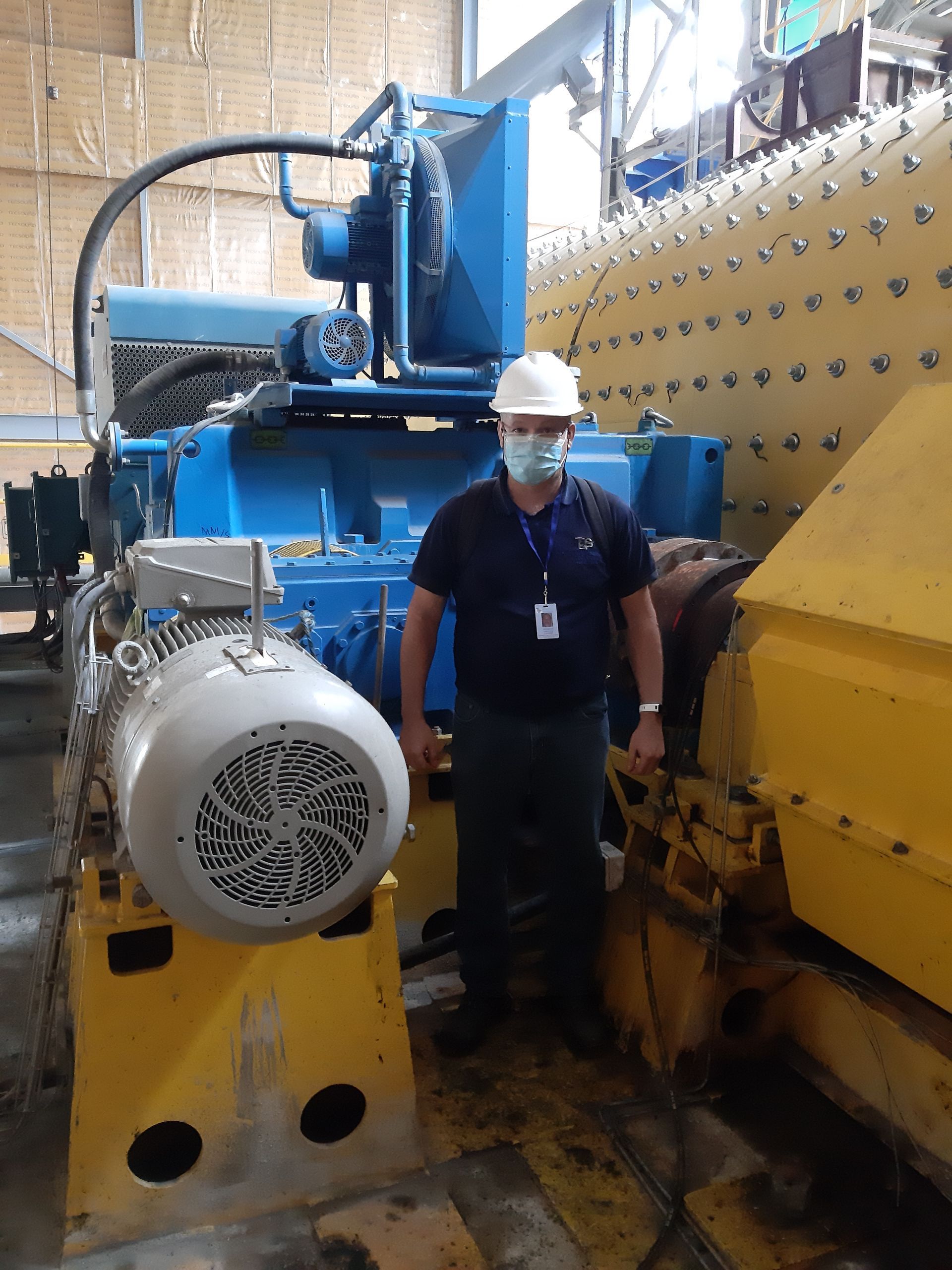 Un hombre con casco y máscara se encuentra frente a una gran maquinaria industrial azul y amarilla en una fábrica.