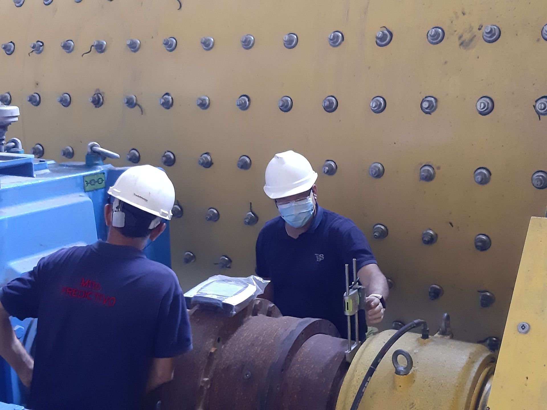 Dos trabajadores con casco inspeccionan maquinaria en una fábrica. Uno sostiene un dispositivo, el otro observa.