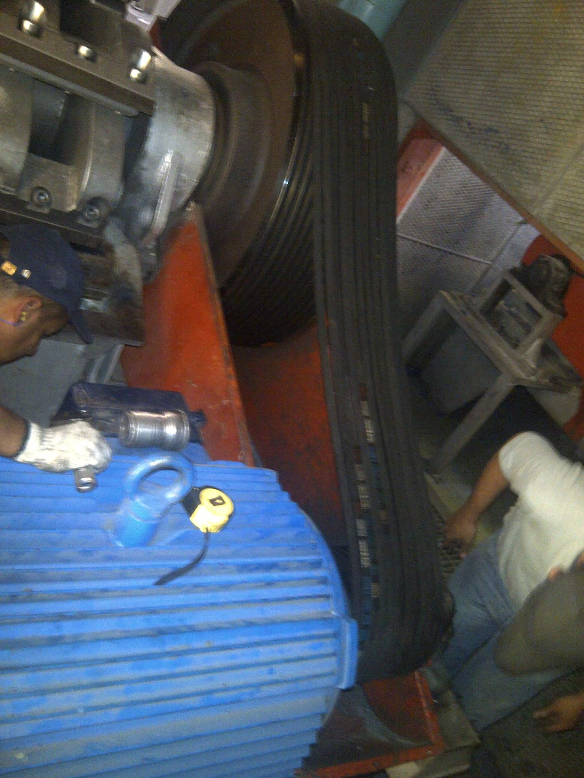 Sistema de transmisión por correa negra y motor azul con dos personas trabajando en él.