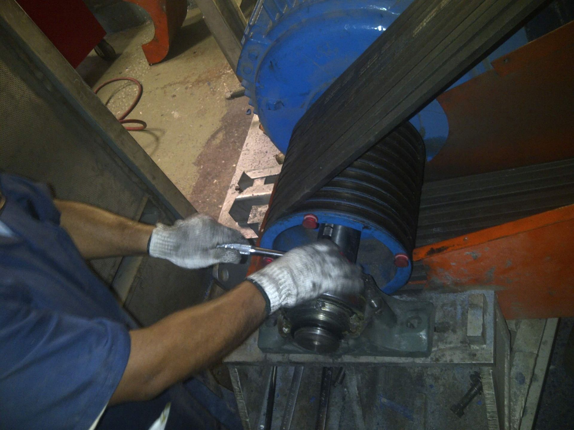 Mecánico con camisa azul y guantes utilizando una llave inglesa en maquinaria con cinturón negro.