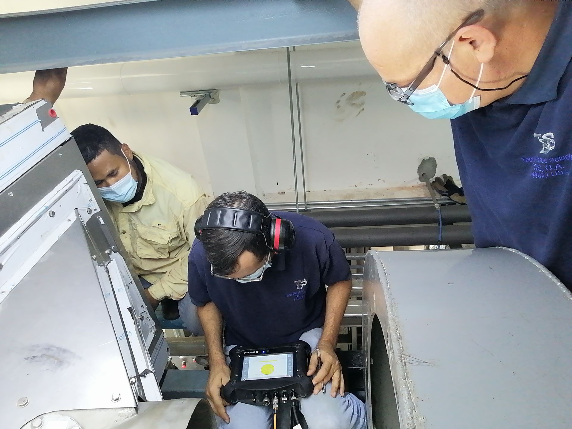 Tres técnicos inspeccionan la maquinaria; uno opera un dispositivo, usando gafas de seguridad y protección para los oídos.