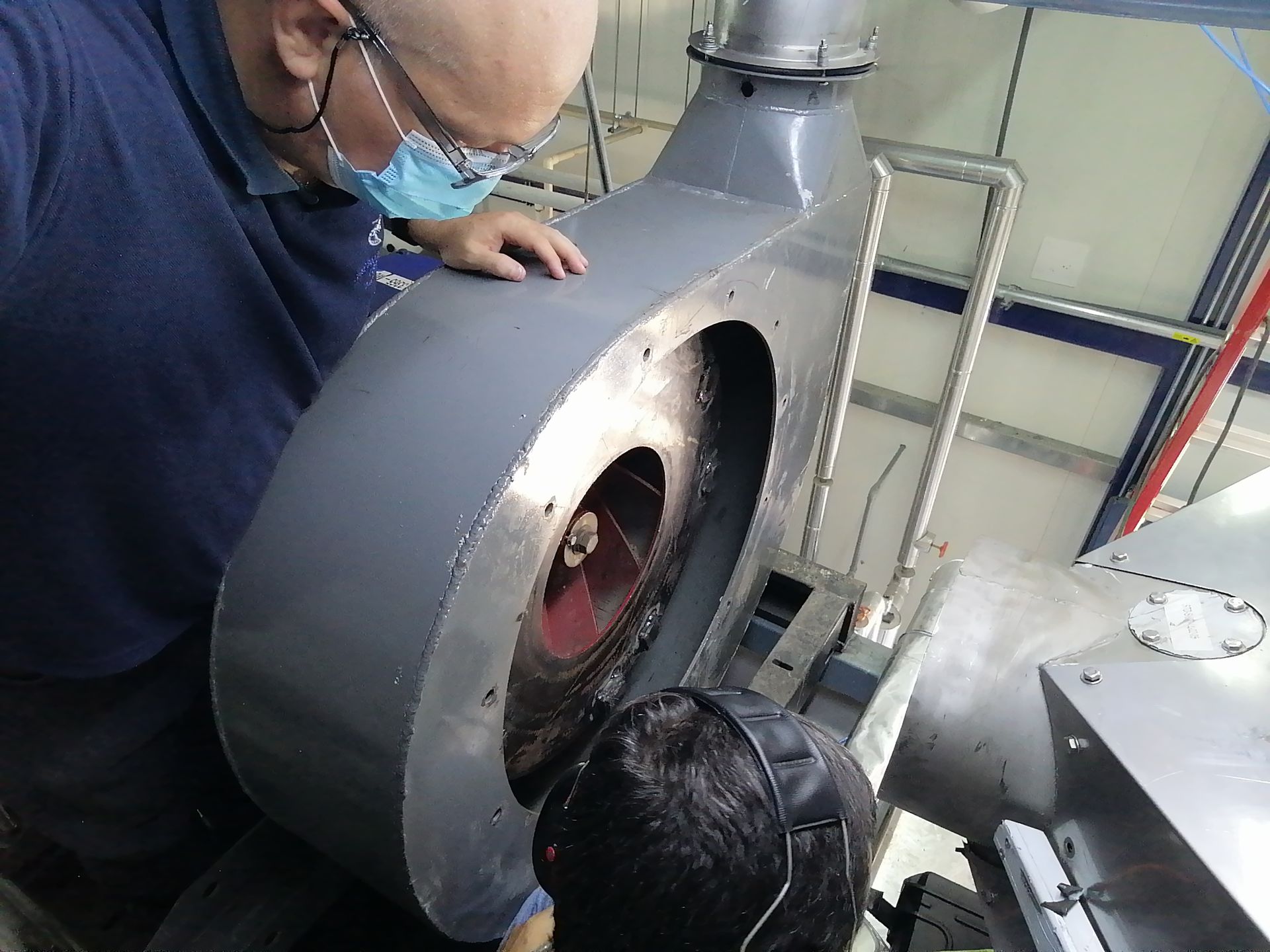 Hombre con mascarilla inspeccionando maquinaria en un taller. Metal gris, primer plano.