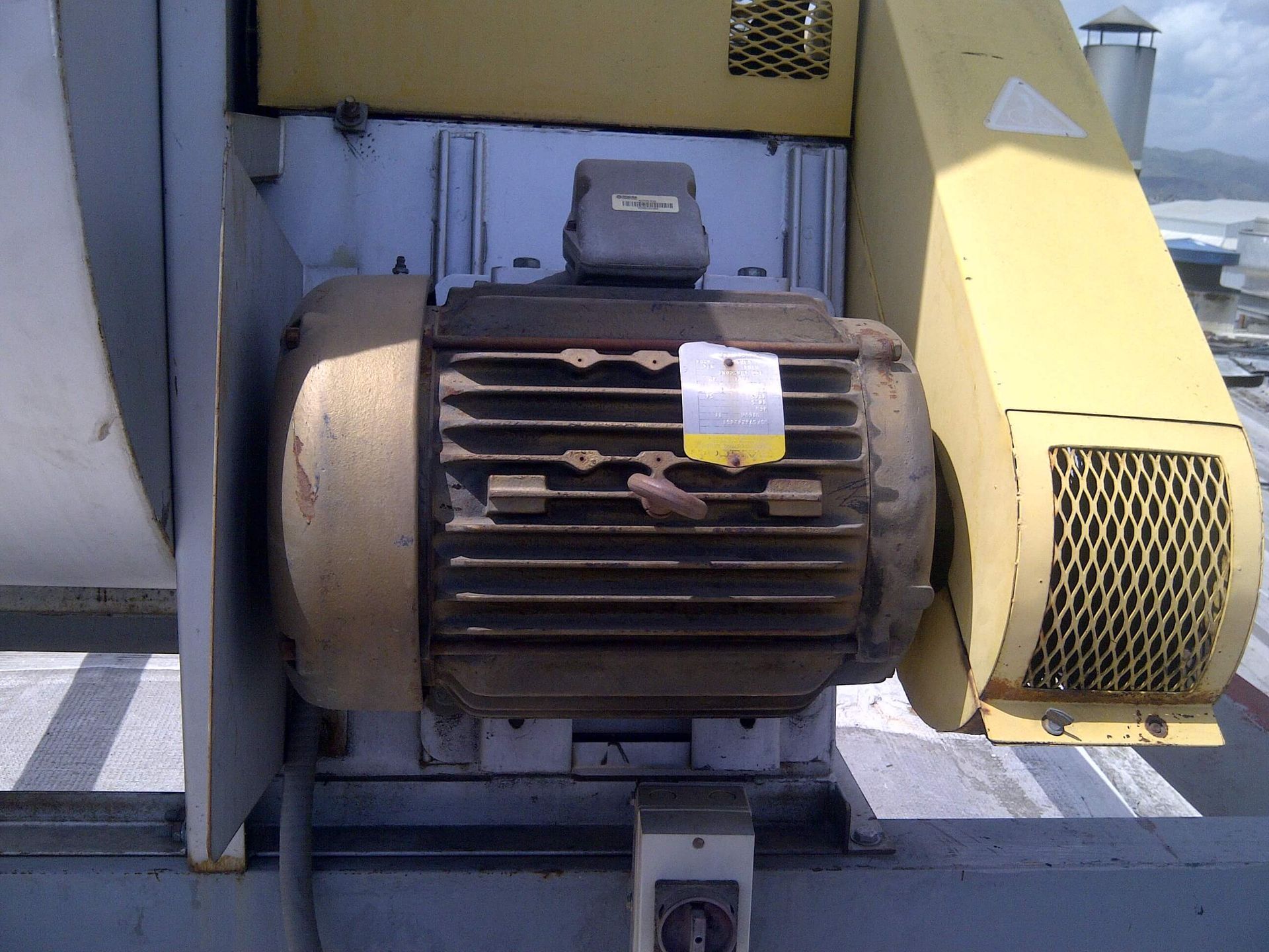 Motor eléctrico, envejecido, con carcasa amarilla, conectado a maquinaria industrial en un tejado.