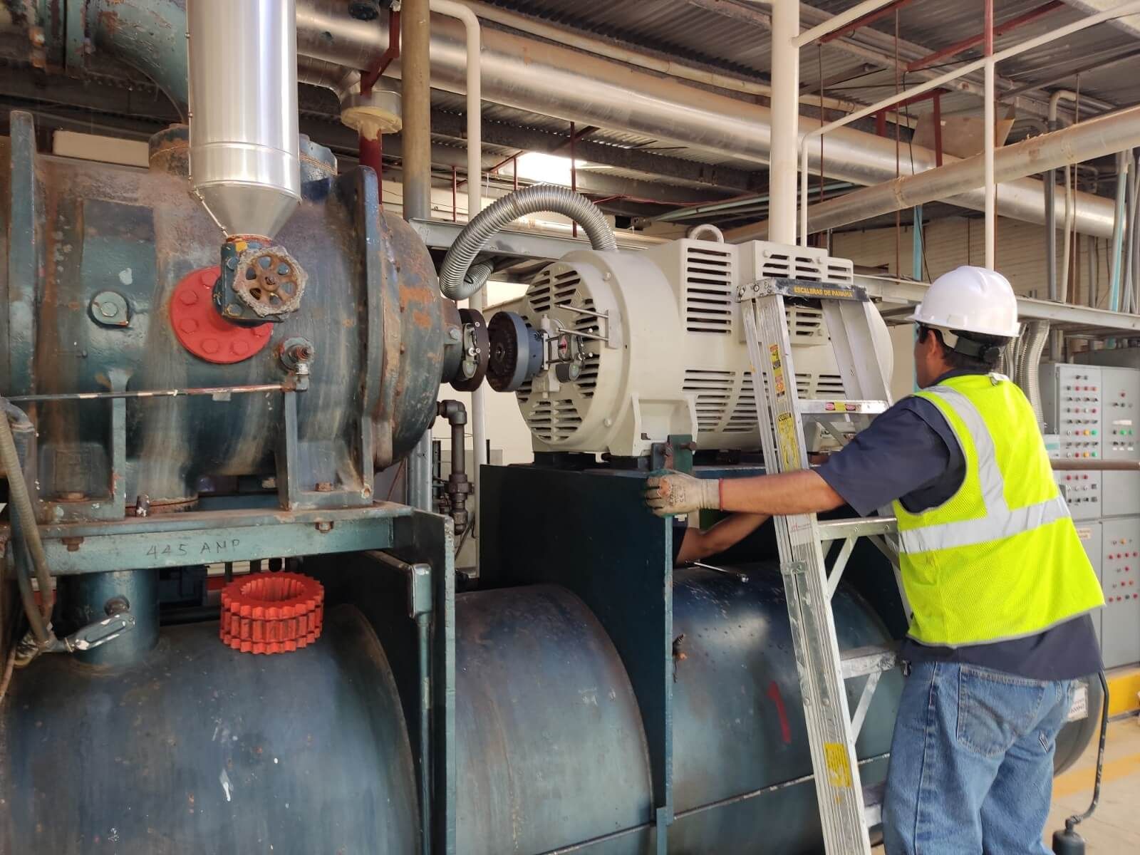Un trabajador con casco y chaleco de seguridad utiliza una escalera para acceder a maquinaria en un entorno industrial.