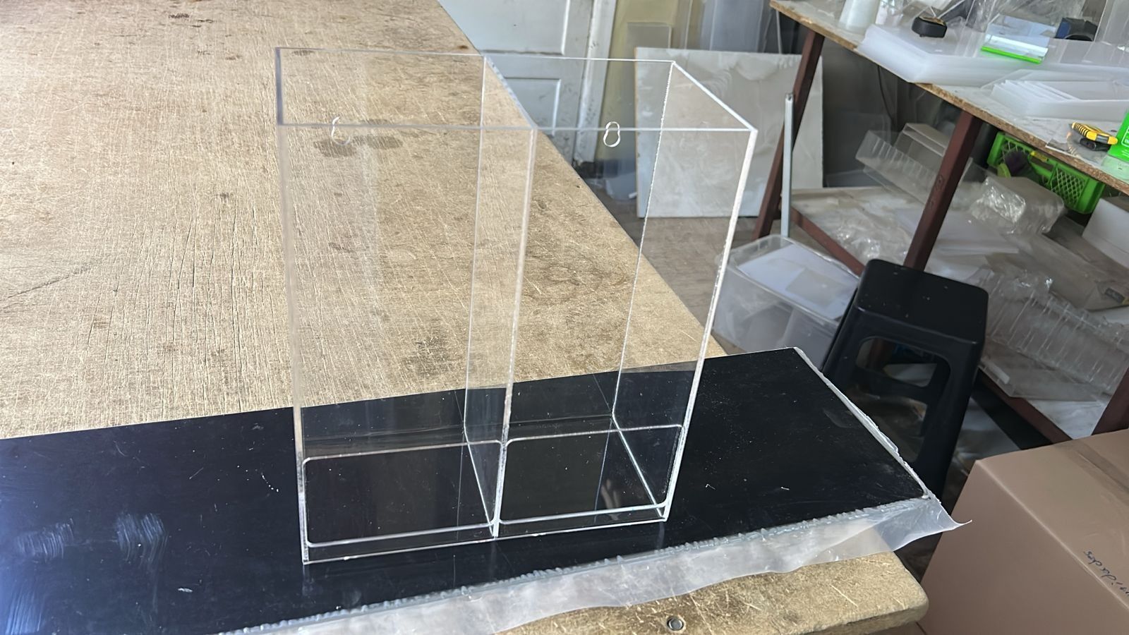 Un recipiente rectangular de acrílico transparente con dos compartimentos, colocado sobre una superficie oscura en un taller.