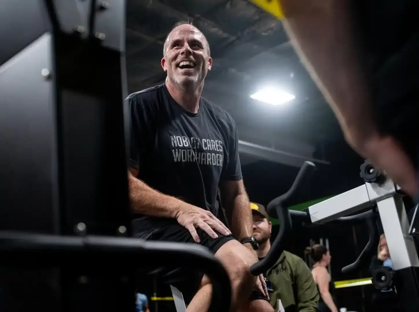 Smiling man in black shirt using gym equipment in a dimly lit fitness room