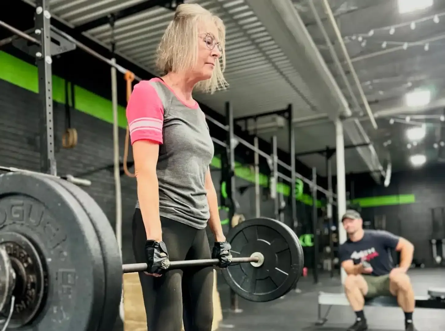 Person lifting a barbell in a gym, with another person exercising in the background.