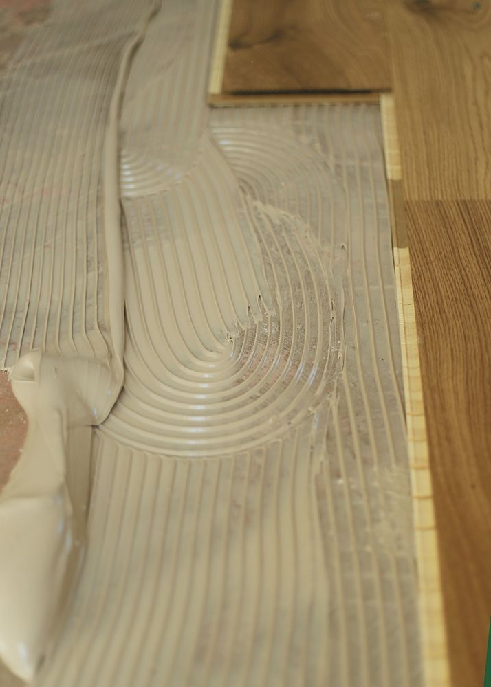 A Person Is Applying Glue To A Wooden Floor — Byron Floor Sanding In Ballina, NSW