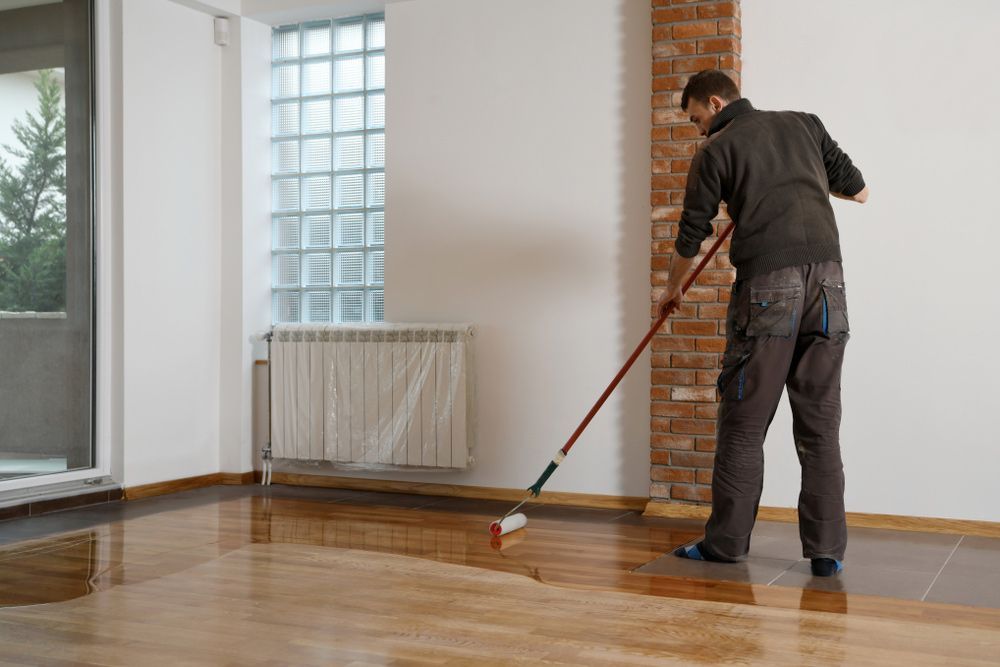 A Man Is Painting A Wooden Floor With A Roller — Byron Floor Sanding In Norther Rivers, NSW