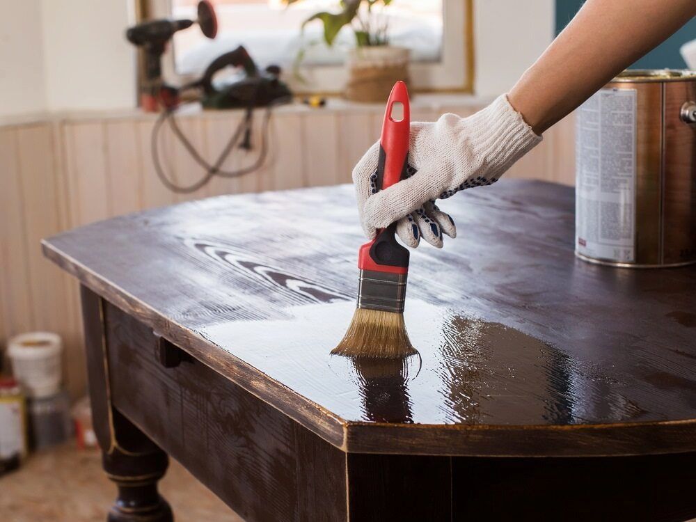 Person Applying Varnish to a Wooden Table With a Brush, Wearing a Glove — Byron Floor Sanding In Tintenbar, NSW