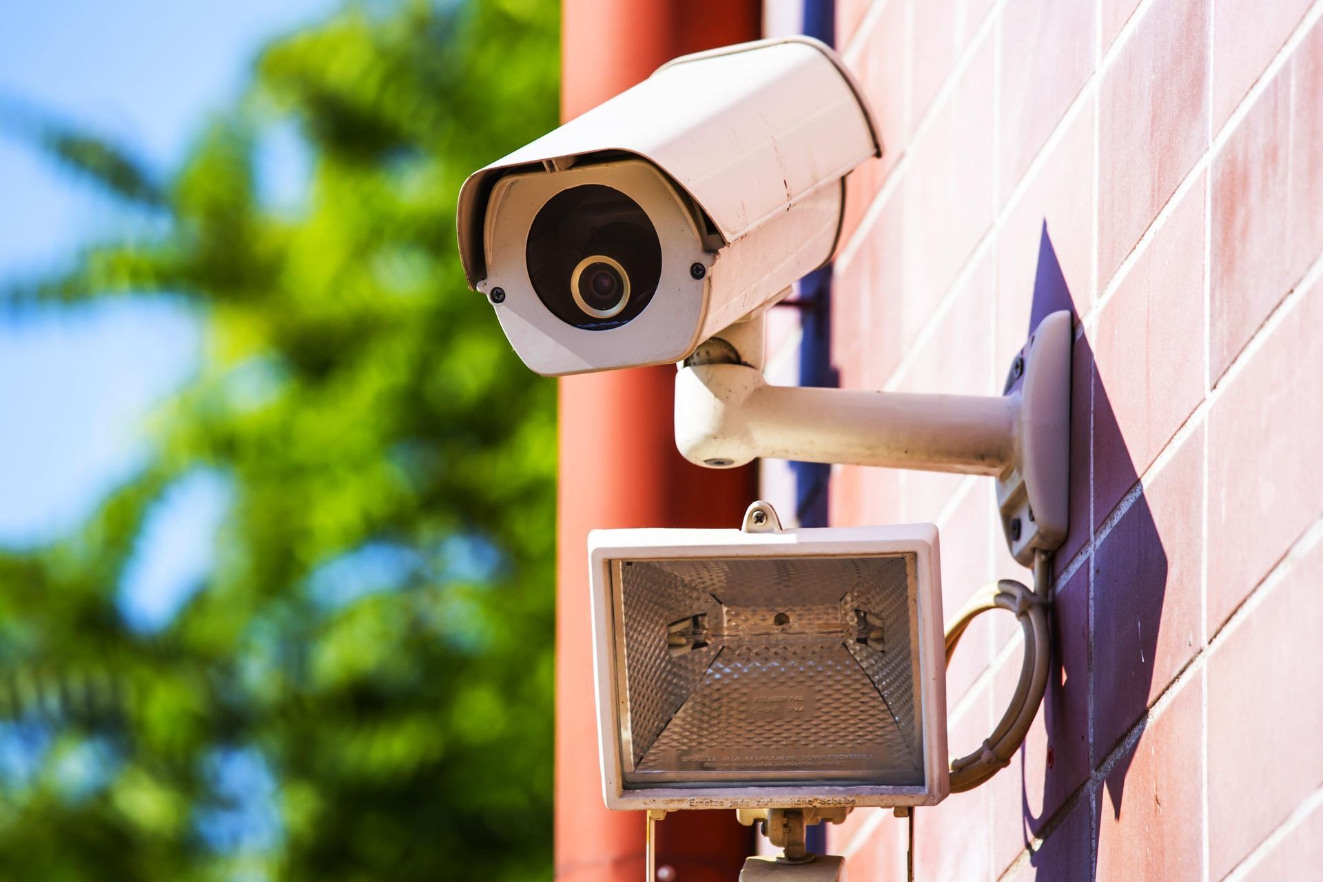 Security camera and floodlight mounted on a red brick wall, with green foliage in the background.